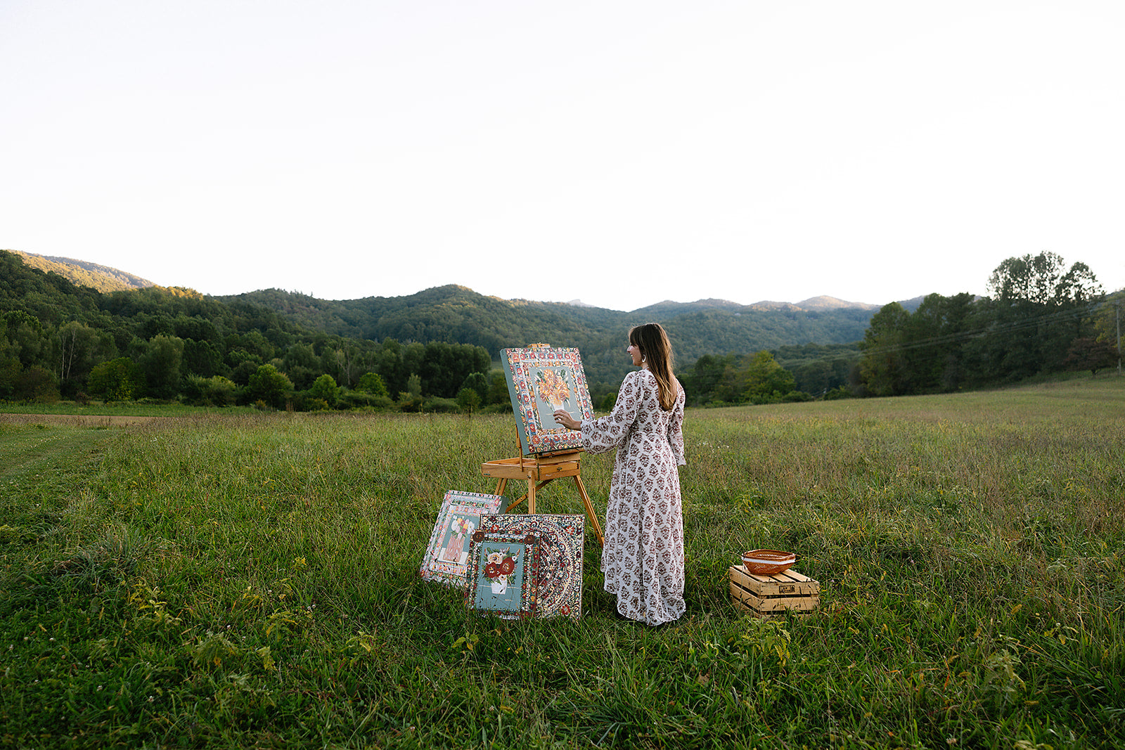 Woman painting outdoors in a scenic field with mountains in the background