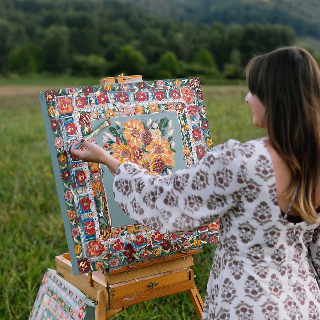 Woman painting a colorful floral scene on an easel in a field with mountains in the background