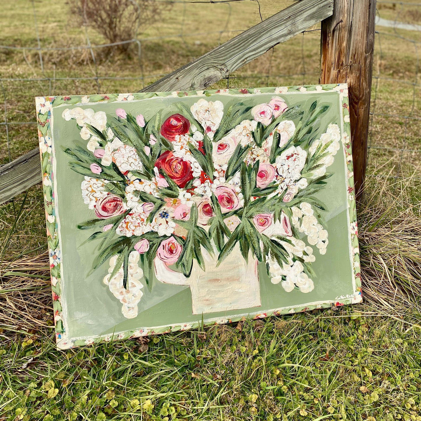 Floral painting on a wooden board in a natural setting with grass and trees.